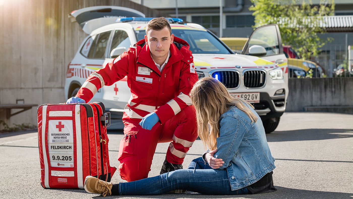 Ein ehrenamtlicher Mitarbeiter in Uniform des Österreichischen Roten Kreuzes hilft einer Frau, die auf einem Parkplatz am Boden sitzt und möglicherweise verletzt ist. Im Hintergrund ist ein Einsatzfahrzeug des Roten Kreuzes zu sehen. Neben dem Ehrenamtlichen ist ein großer roter Rucksack des Roten Kreuzes zu sehen.